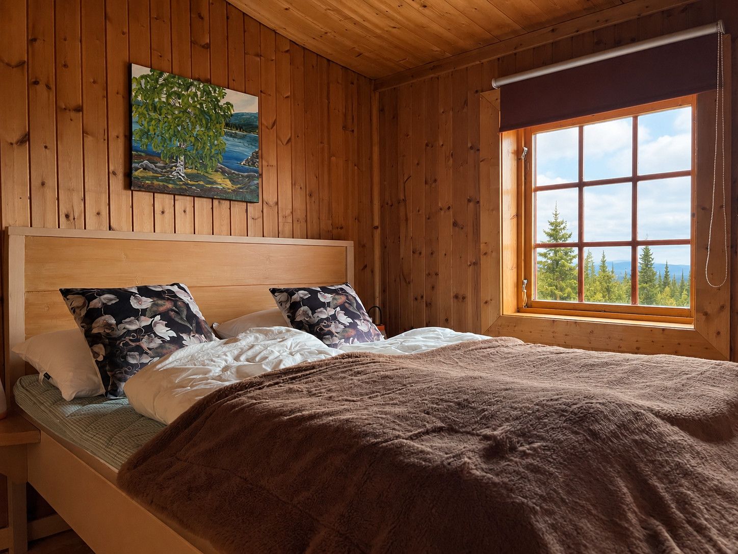 Een slaapkamer in een houten hutje, met een bed, kussens, een deken, een schilderij en een raam met uitzicht.