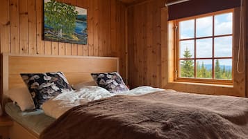 Een slaapkamer in een houten hutje, met een bed, kussens, een deken, een schilderij en een raam met uitzicht.