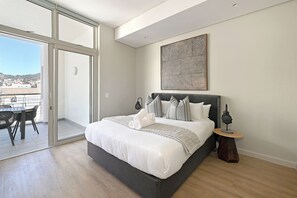A bedroom with a large bed, a view through a sliding glass door, a painting on the wall, and a small table with a lamp. - Cape Town City 2-Bed, Views, Balcony & Pool (Cape Town)