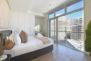 Un dormitorio moderno con una cama grande, una lámpara de noche, un espejo y vistas de la ciudad a través de amplias ventanas.