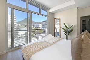 A bedroom with a large window, a bed with a beige blanket, a mirror, and a plant. - Cape Town City 2Bed, Views, Balcony, Pool (Cape Town)
