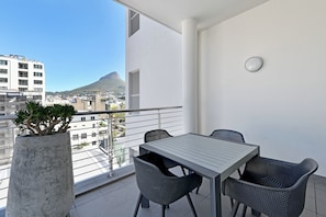Un balcón con mesa y sillas, una planta en maceta y vista a un paisaje urbano con una montaña.