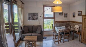Ein gemütliches Wohnzimmer mit einer Couch, einem kleinen Tisch, einem Esstisch mit Stühlen, einer Lampe und einem Fenster mit Blick auf Grünflächen.