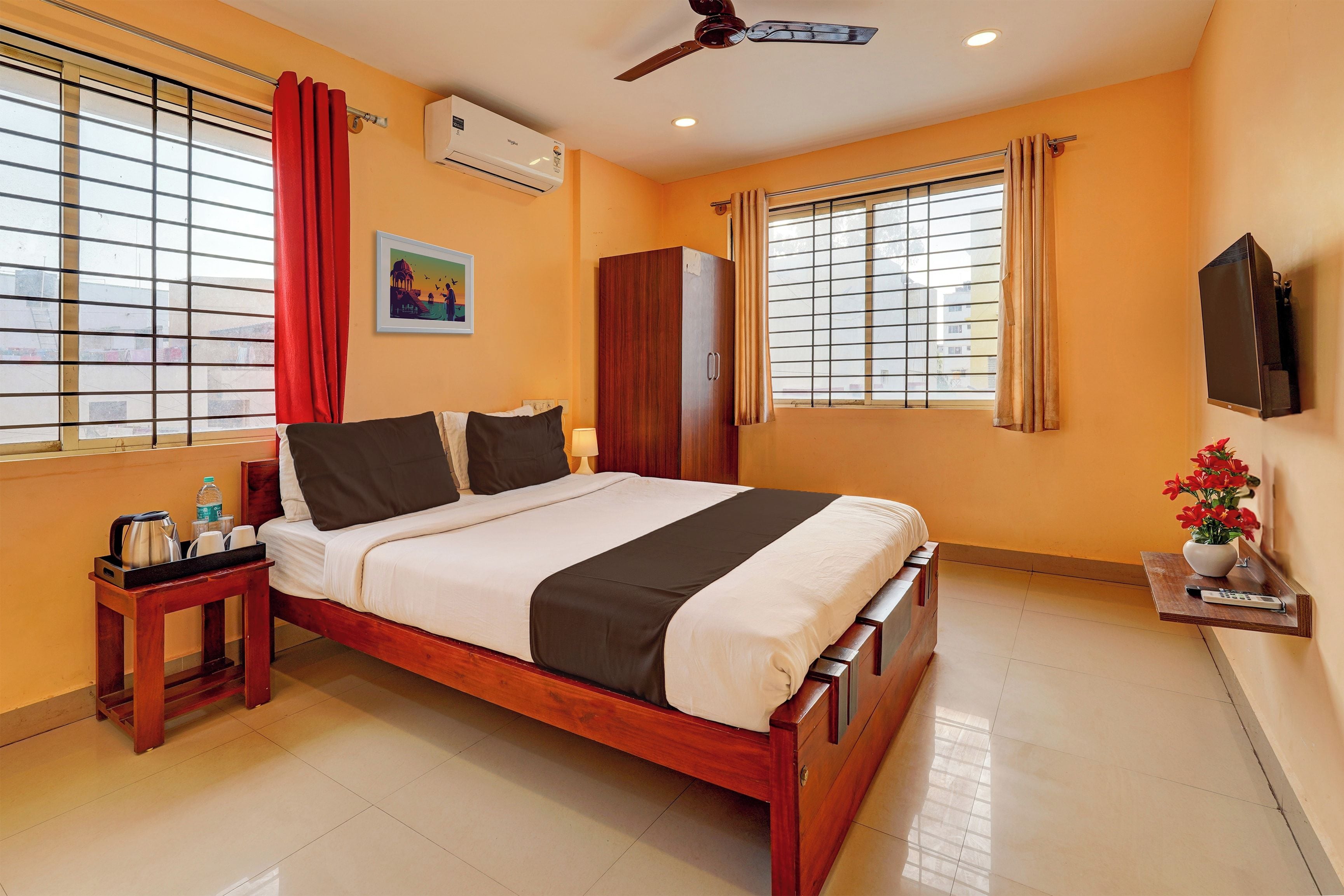Habitación de hotel con cama, armario, mesita de noche con hervidor y botellas, televisor montado en la pared y ventana con cortinas.