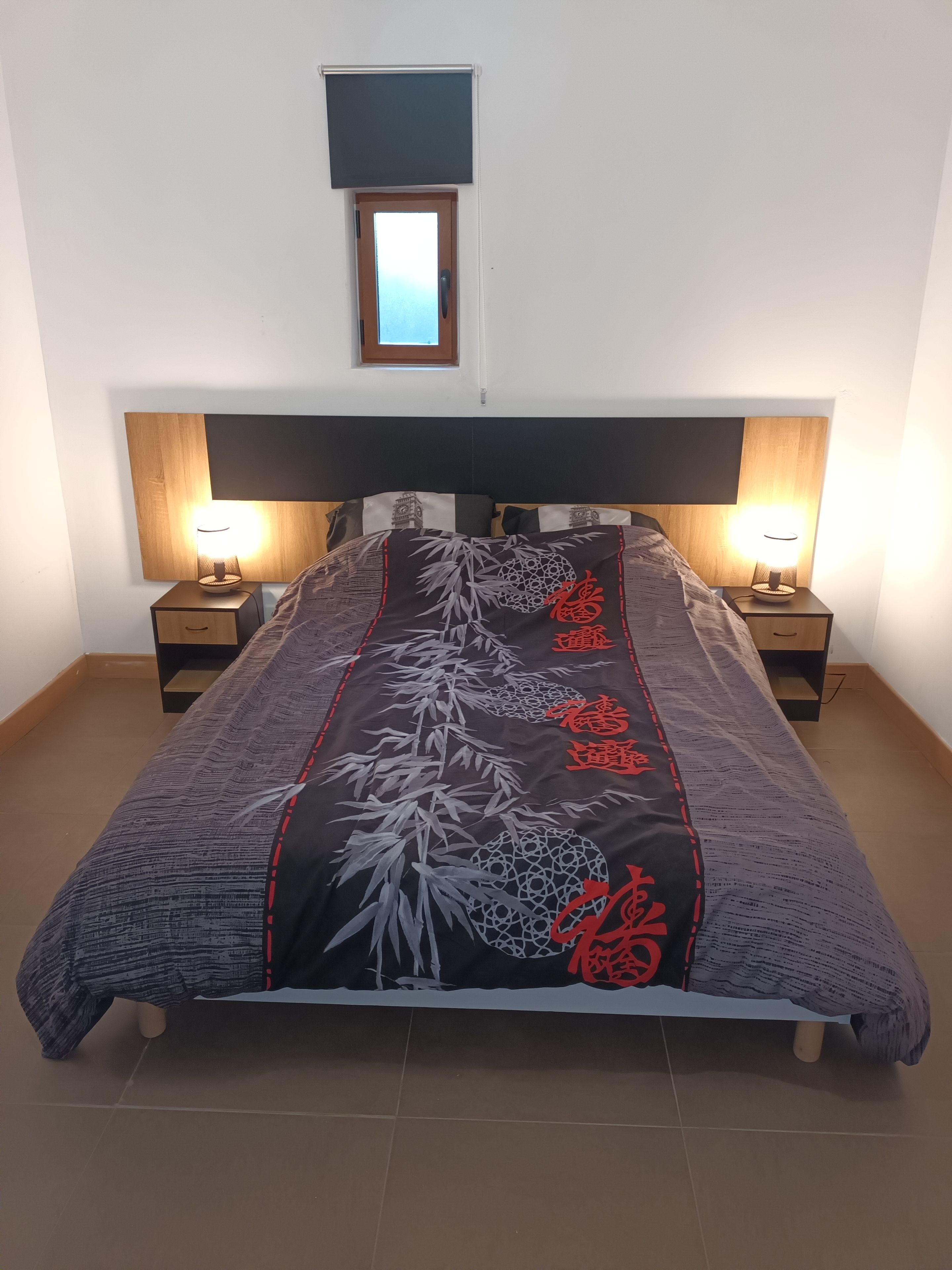 Un lit avec une couvre-lit à motifs, deux tables de chevet avec des lampes, un téléviseur fixé au mur et une petite fenêtre.