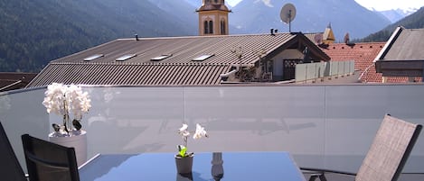 A dining area with a table and chairs, a view of mountains, and a church with a clock tower.
