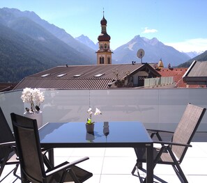 A dining area with a table and chairs, a view of mountains, and a church with a clock tower. - Deluxe Apartment in the Stubai Valley—where high standards meet cozy comfort (Fulpmes)