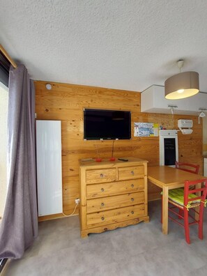 Ein Zimmer mit einer Holzwand, einem Flachbildfernseher, der über einer Holzkommode angebracht ist, einem Esstisch mit Stühlen und einer Deckenleuchte.