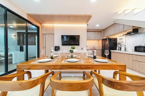 A modern kitchen with wooden cabinetry, a dining table set for four, and a flat-screen TV mounted on the wall. - Serene Pool Villa Pattaya 2 (Chonburi)