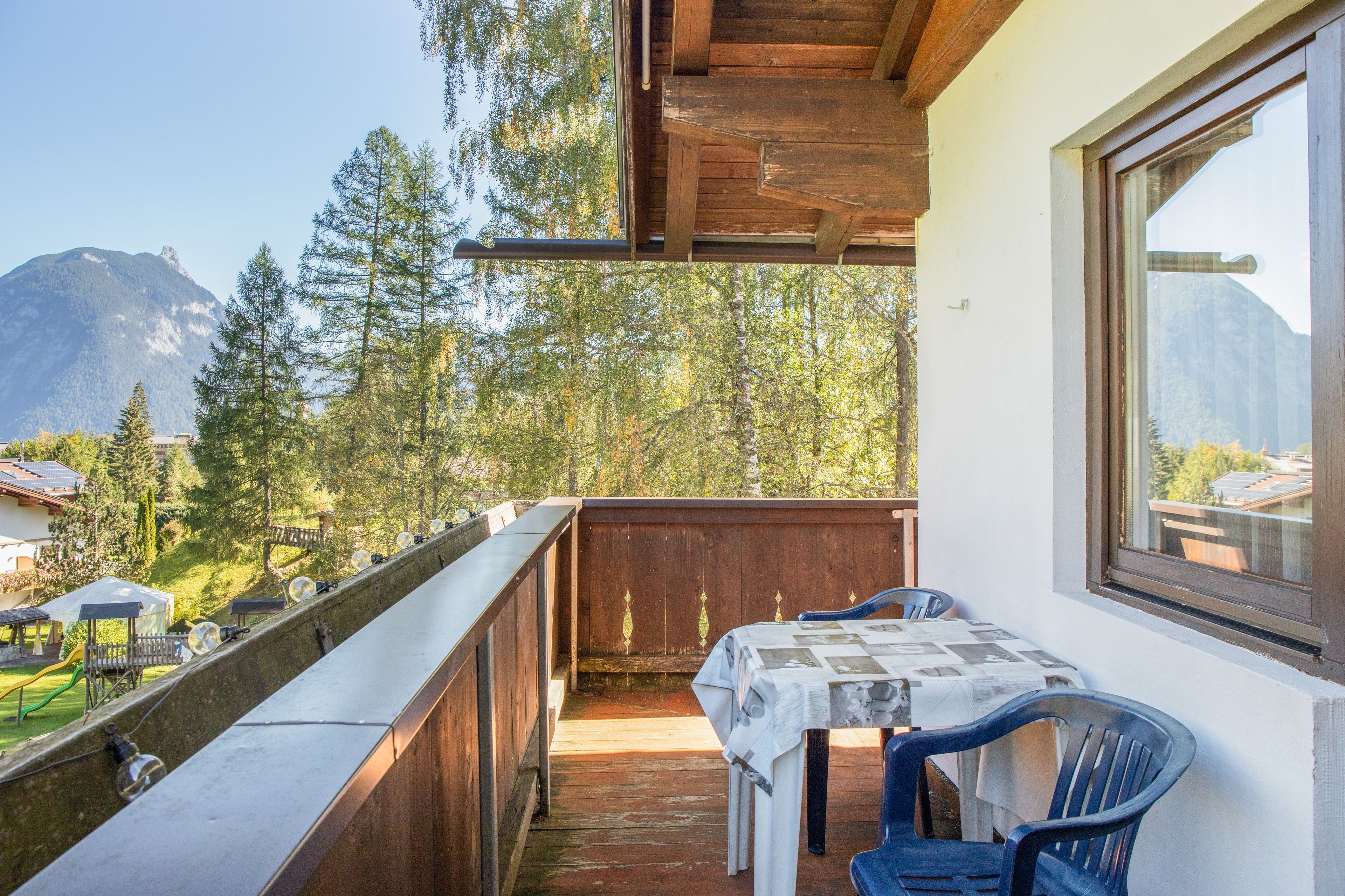 Un balcón con mesa y sillas, con vistas a un paisaje montañoso con árboles y edificios.