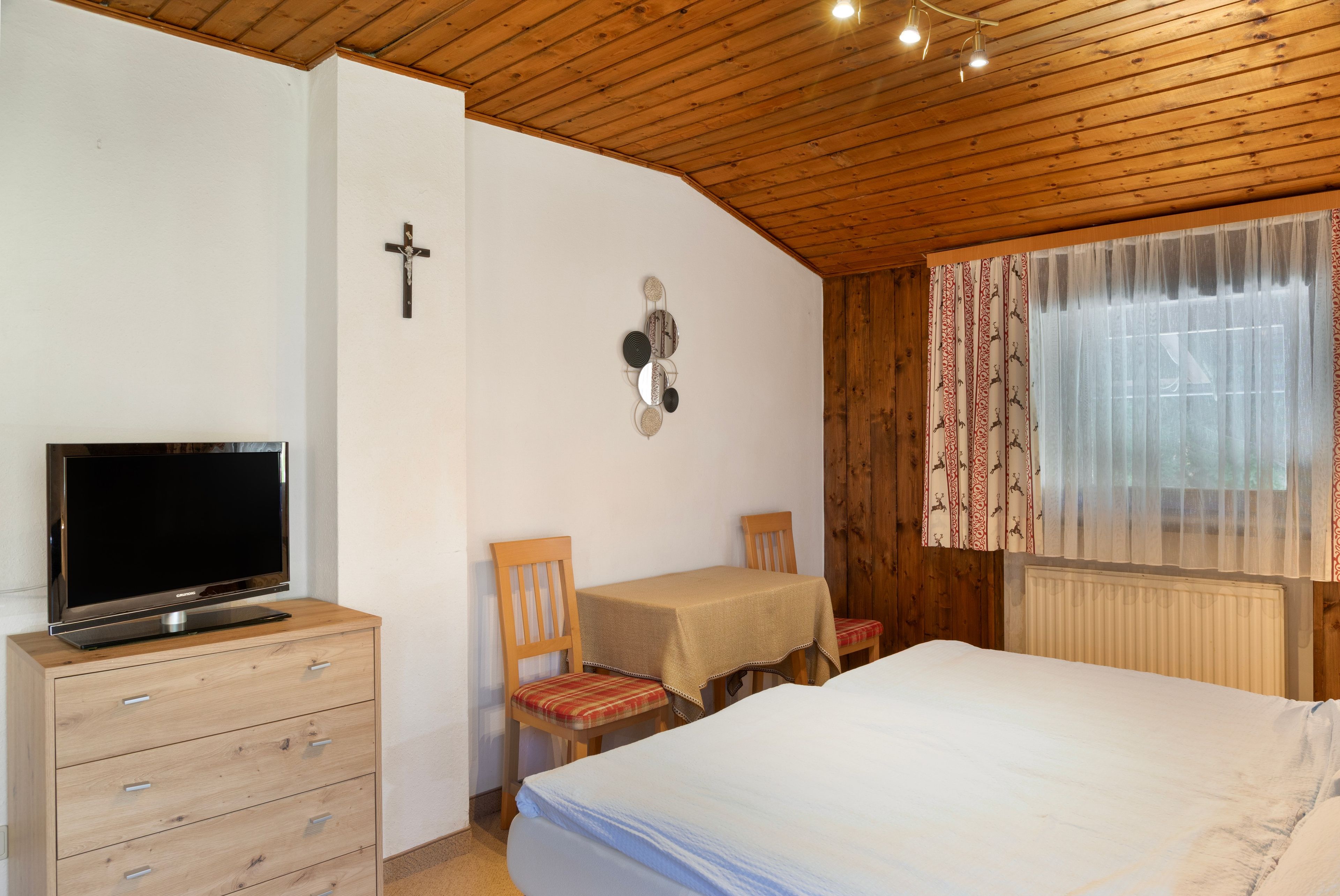 Een slaapkamer met een bed, een tv op een dressoir, een kruis aan de muur en een raam met gordijnen.