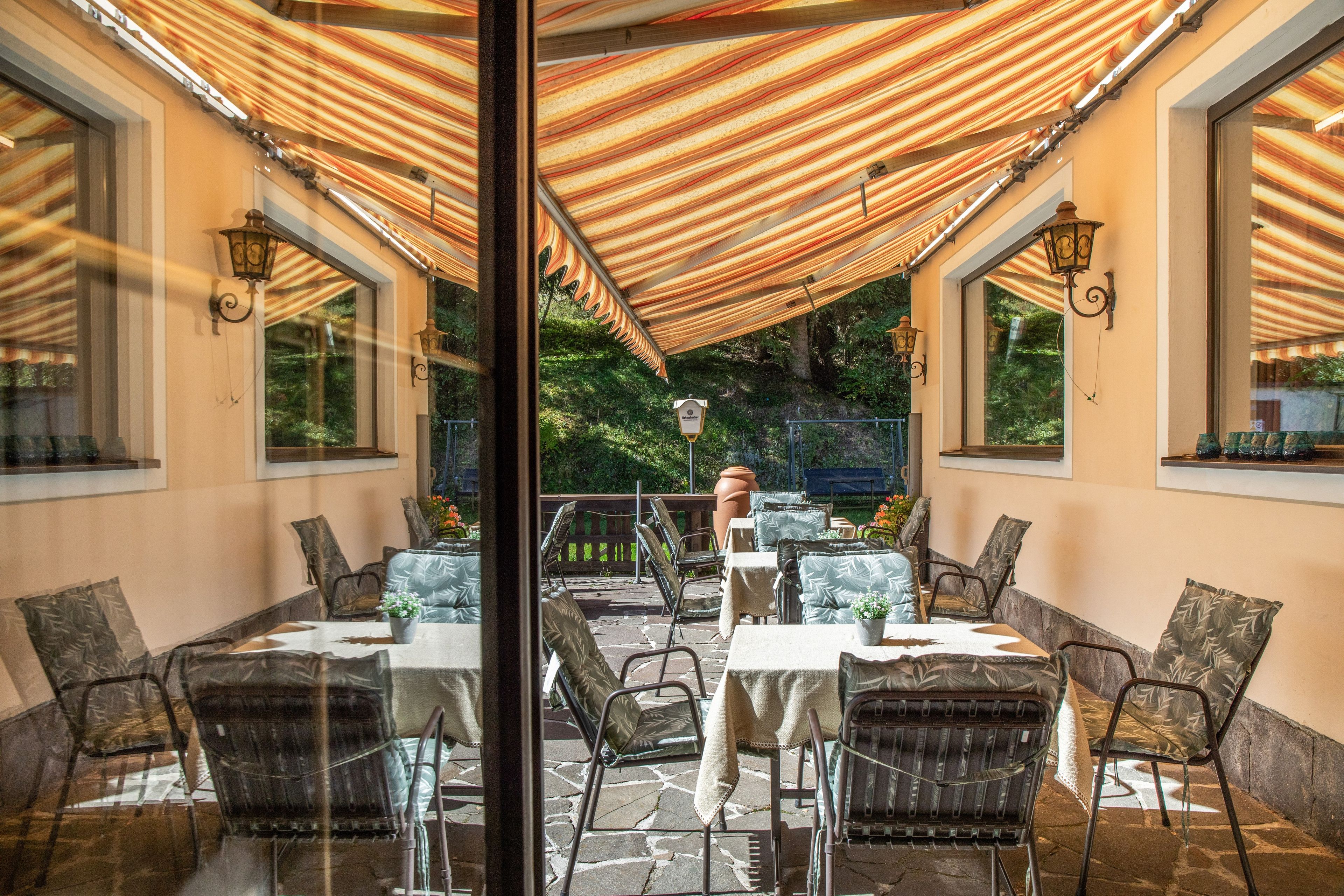 Een terras buiten met tafels en stoelen, een pergola erboven en uitzicht op groen.