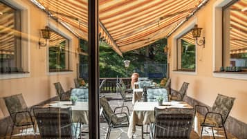 Een terras buiten met tafels en stoelen, een pergola erboven en uitzicht op groen.