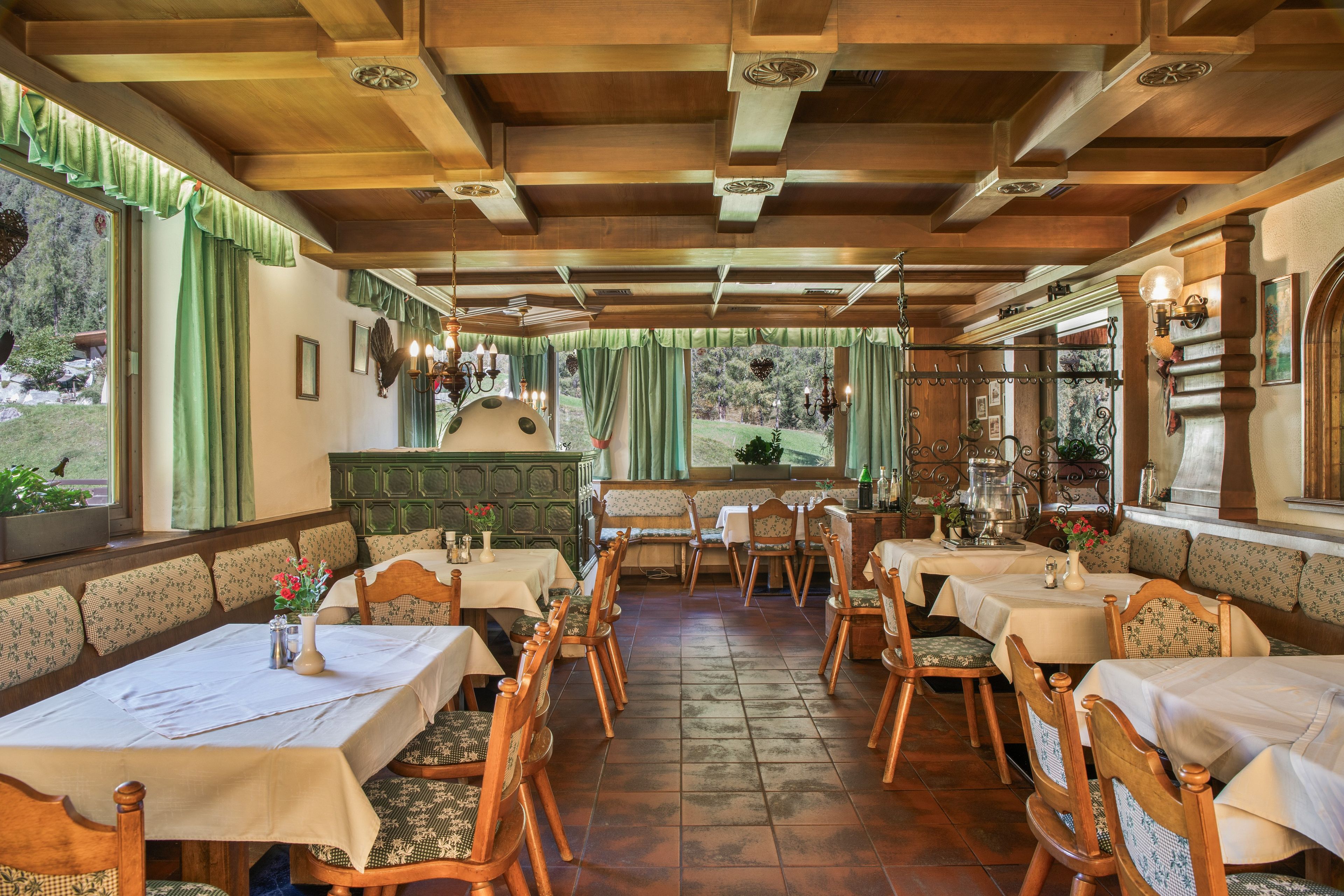 Een restaurant met houten balken, groene gordijnen en uitzicht op het landschap. Tafels zijn gedekt met witte tafelkleden en houten stoelen.