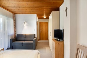 Een kamer met een houten plafond, een grijze bank, een televisie op een houten dressoir, en een deur naar een andere kamer.