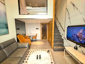 A modern living room with a sofa, a coffee table with remotes, a TV, and a staircase leading to an upper level. - Huanyue Serviced Apartments  (Shenzhen)