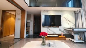 A modern living room with a flat-screen TV mounted on a marble wall, a wooden cabinet, a vase with red flowers, and a scale on wheels. - Huanyue Serviced Apartments  (Shenzhen)