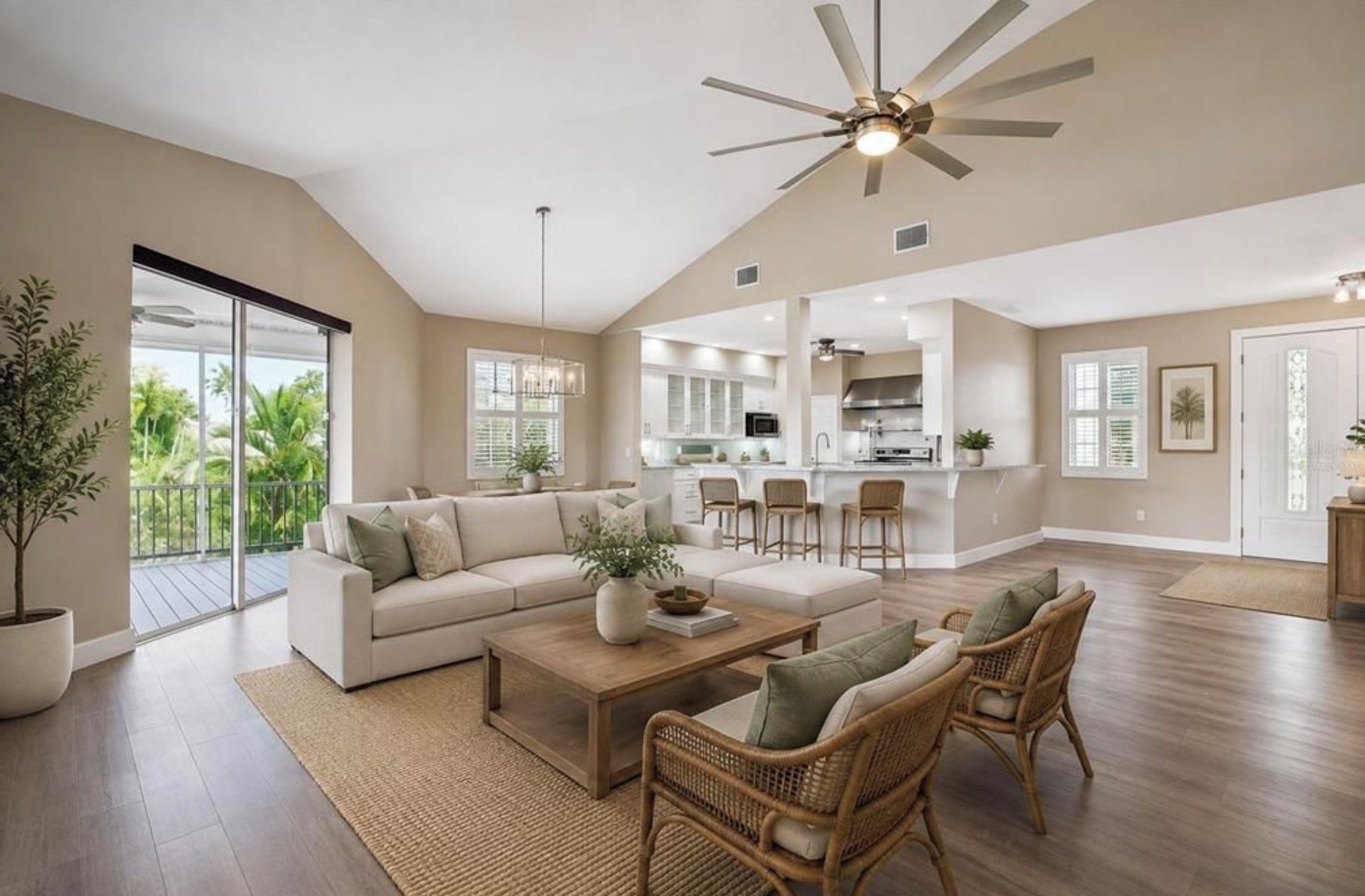 A spacious living area with a large sofa, a coffee table, and wicker chairs. There is a ceiling fan, a kitchen in the background, and a view of the outdoors through large windows.