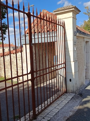 A gated entrance with a metal fence, a stone wall, and a building with a tiled roof. - In the heart of town, duplex with parking and secure bike storage. (Fontenay-le-Comte)