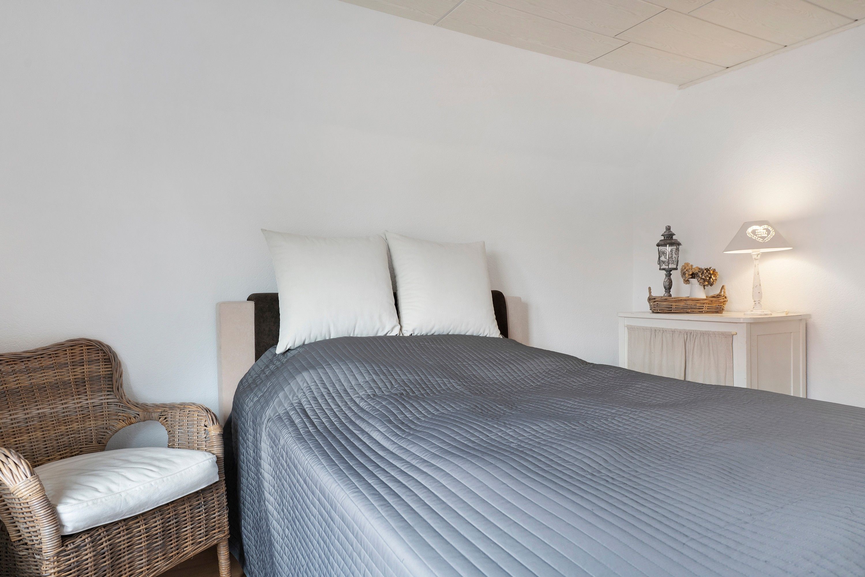 Ein Schlafzimmer mit einem Bett, einem Korbsessel, einem Nachttisch mit Lampe und einem an der Wand befestigten Regal.