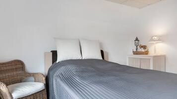 Ein Schlafzimmer mit einem Bett, einem Korbsessel, einem Nachttisch mit Lampe und einem an der Wand befestigten Regal.