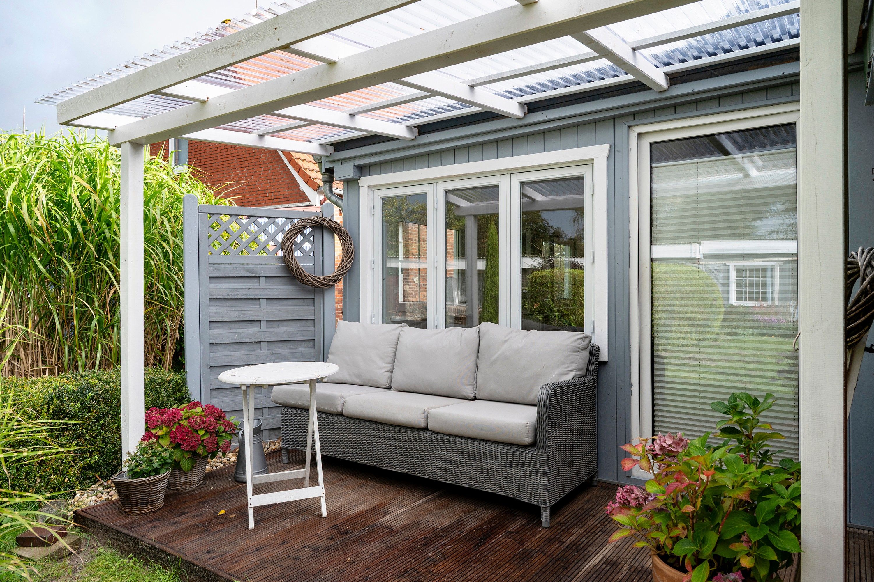 Une terrasse couverte avec un canapé et une table, entourée de plantes et d’un mur en briques.