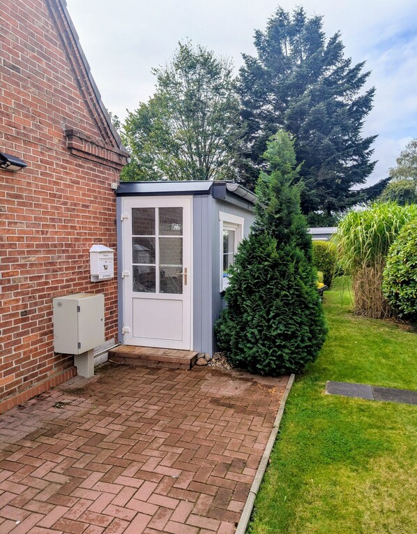 A brick house with a small shed, a well-maintained lawn, and a tall evergreen tree. - Holiday Home 'Lilly' with Private Terrace, Private Garden and Wi-Fi (Meldorf)