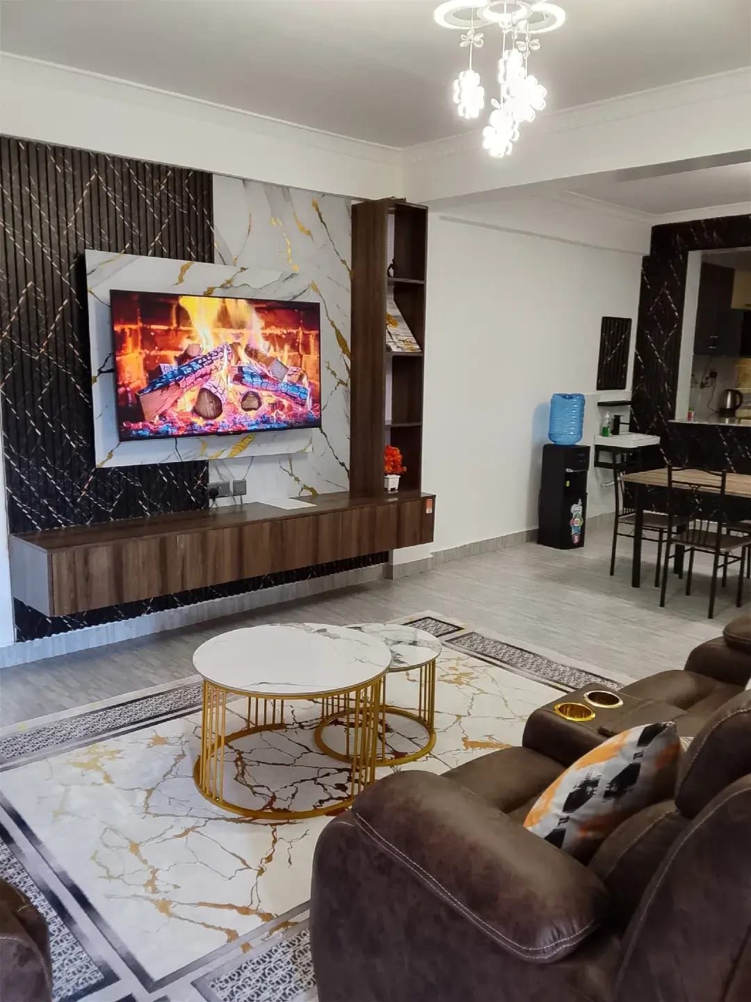Una sala moderna con un televisor de pantalla plana montado en una pared de mármol, un sofá de cuero marrón, una mesa de centro redonda y un comedor al fondo.
