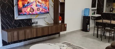 Una sala moderna con un televisor de pantalla plana montado en una pared de mármol, un sofá de cuero marrón, una mesa de centro redonda y un comedor al fondo.