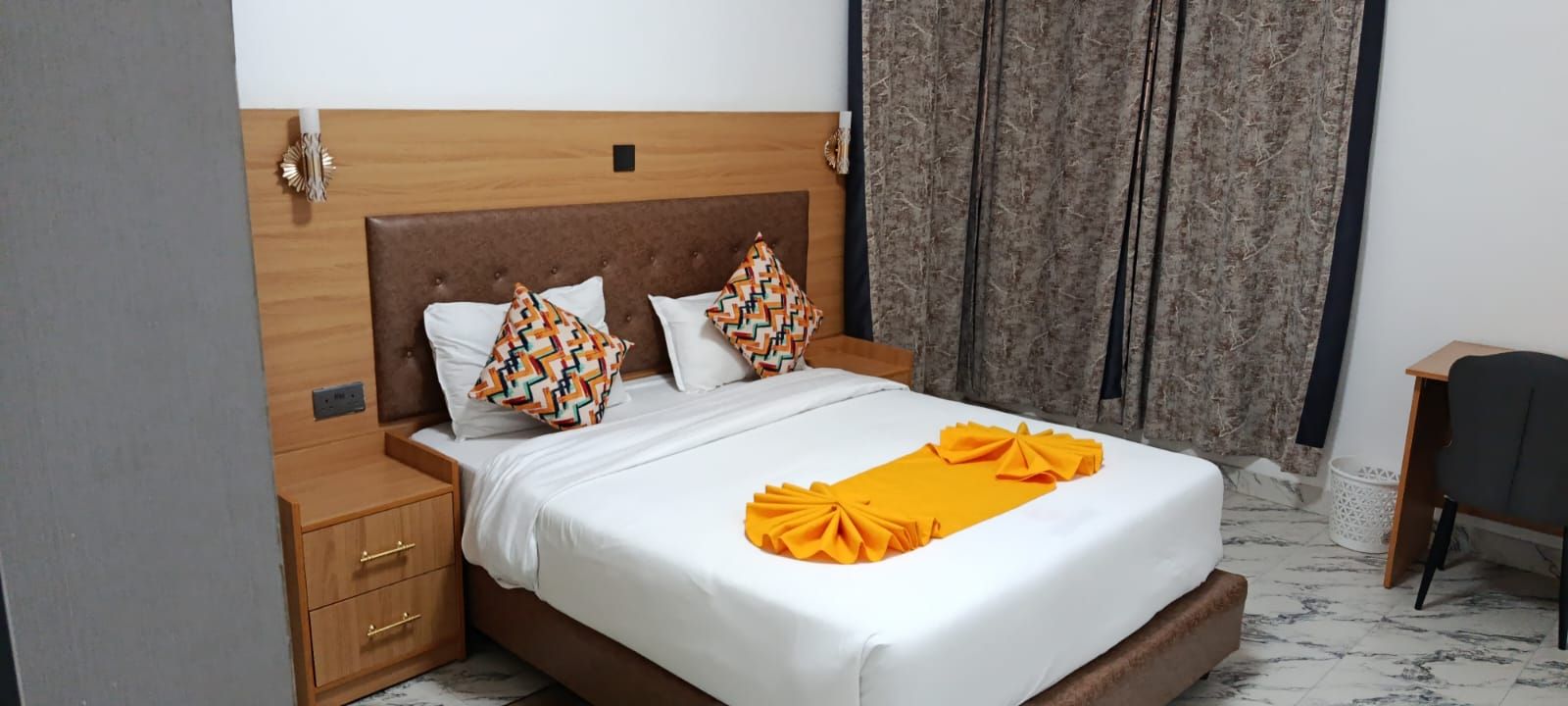 Una cama bien tendida con una colcha blanca y un lazo decorativo naranja, una mesita de noche con cajones, un cabecero con panel de madera y una cortina con estampado de hojas.