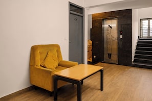 Een kamer met een gele fauteuil, een houten tafel, een grijze deur en een trap met een lift.