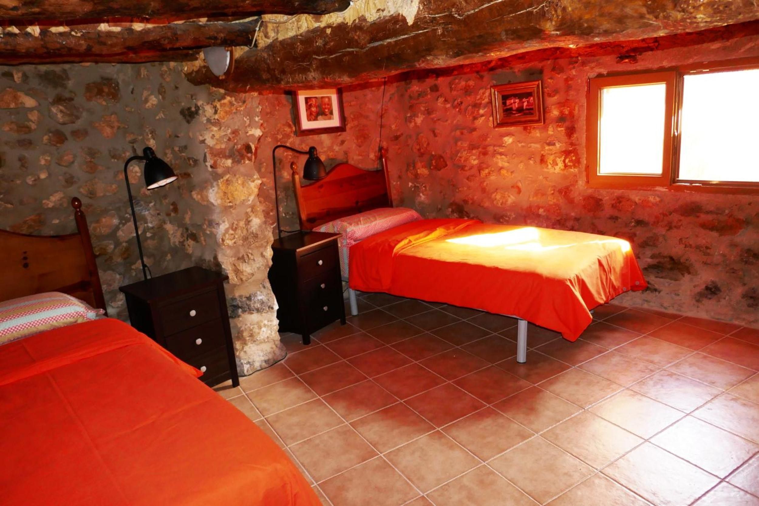 Un dormitorio con pared de piedra rústica, una cama individual con cubrecama naranja, una mesita de noche con lámpara y una ventana con luz solar.