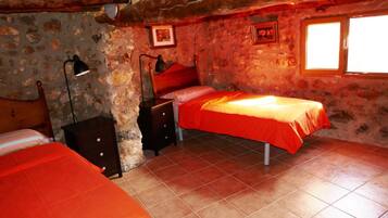 Un dormitorio con pared de piedra rústica, una cama individual con cubrecama naranja, una mesita de noche con lámpara y una ventana con luz solar.