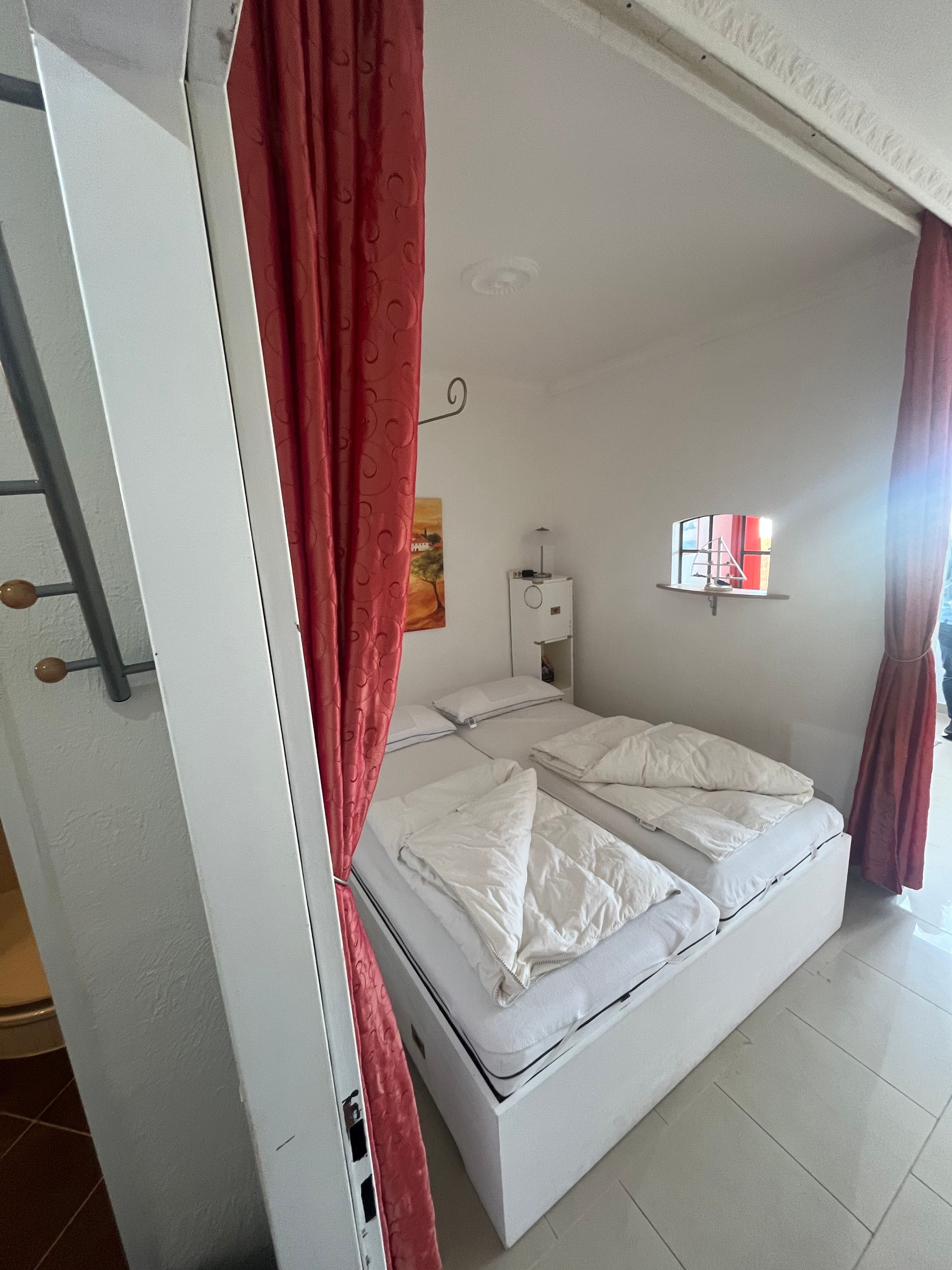 Ein Zimmer mit einem Einzelbett, einer weißen Matratze, einem kleinen Nachttisch mit Lampe, einem Wandregal und einem Fenster mit Vorhängen.
