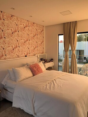 A bedroom with a large bed, a headboard with a flamingo pattern, a bedside table, and a view of the outdoors through a window with curtains. - Celias Beautiful House (Tibau do Sul)