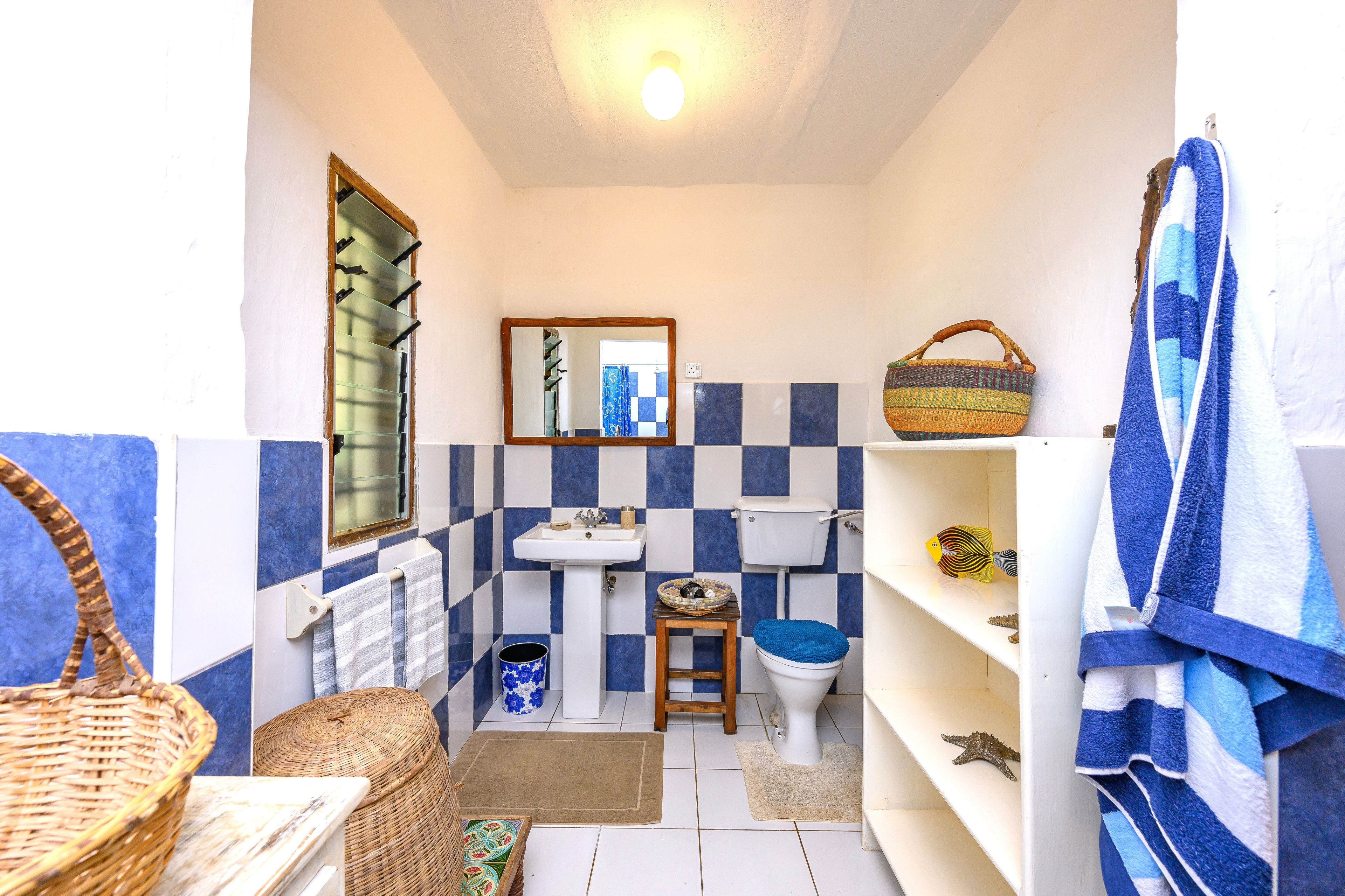 Une petite salle de bain avec un lavabo, des toilettes et un miroir. On y trouve des serviettes et un panier sur les étagères.