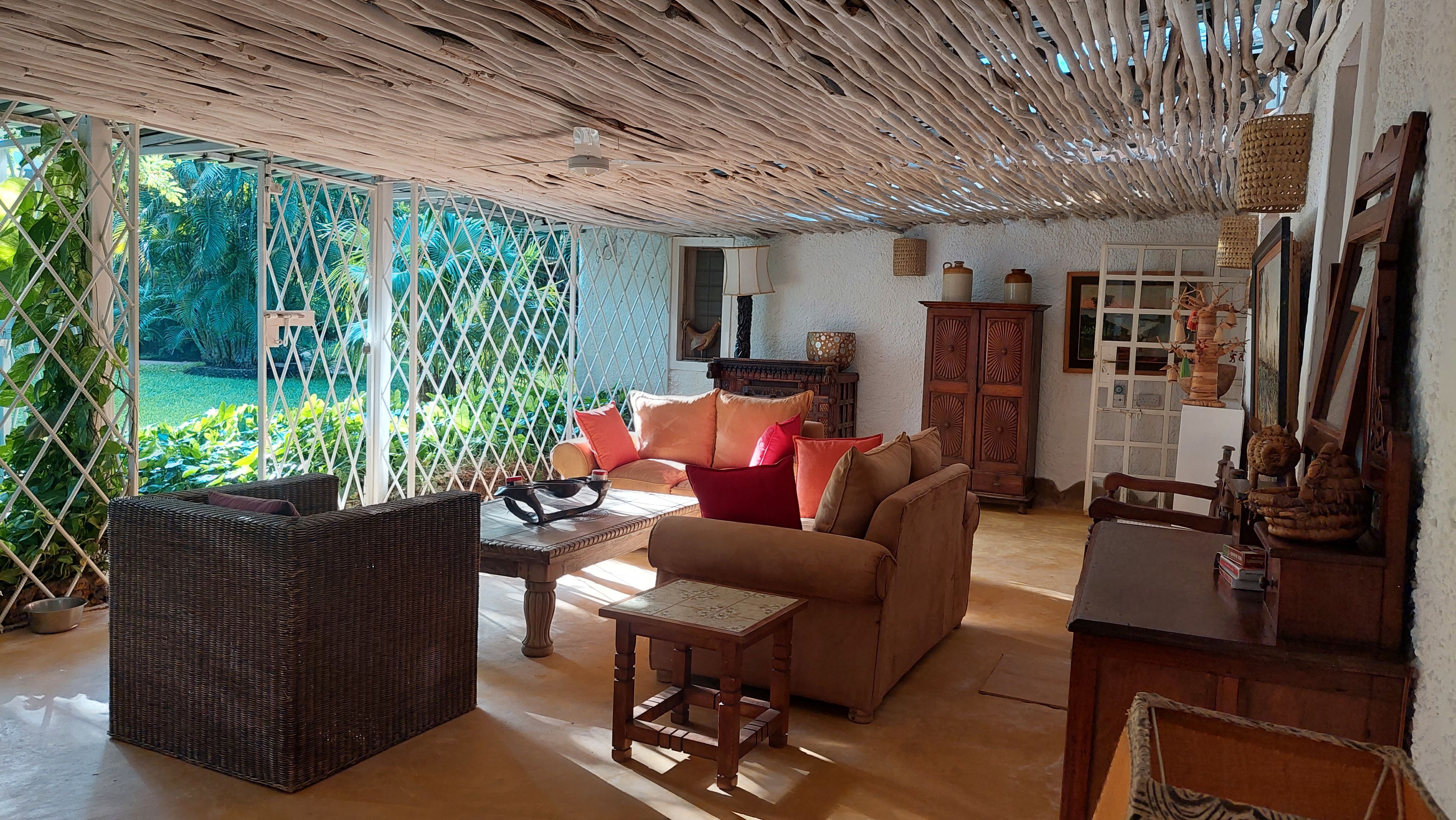 A cozy living room with wicker furniture, a wooden cabinet, and a view of a lush garden through a lattice window.