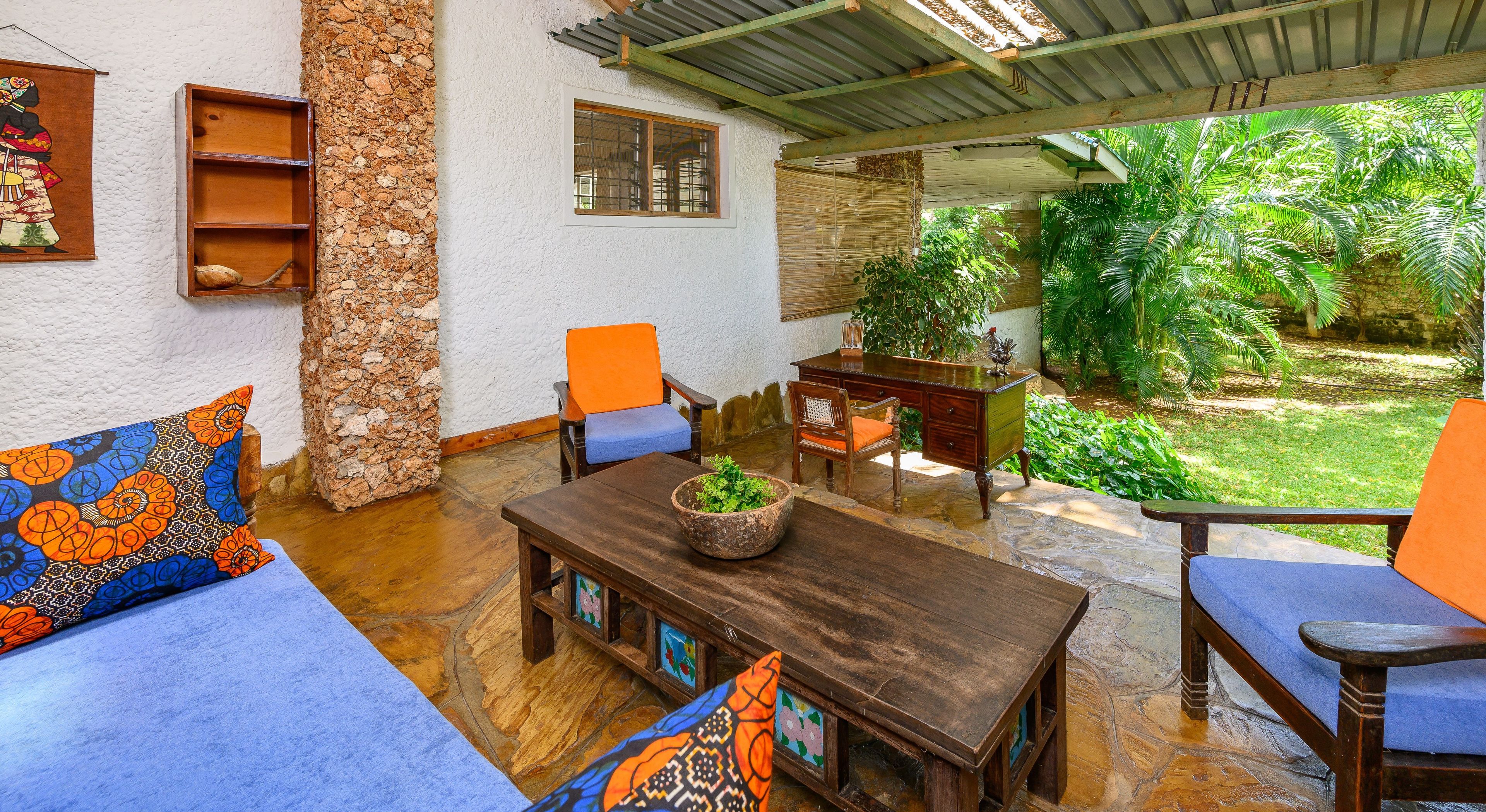 Un patio techado con pared de piedra, una mesa de madera con un cuenco de plantas y un sofá azul con cojines coloridos.