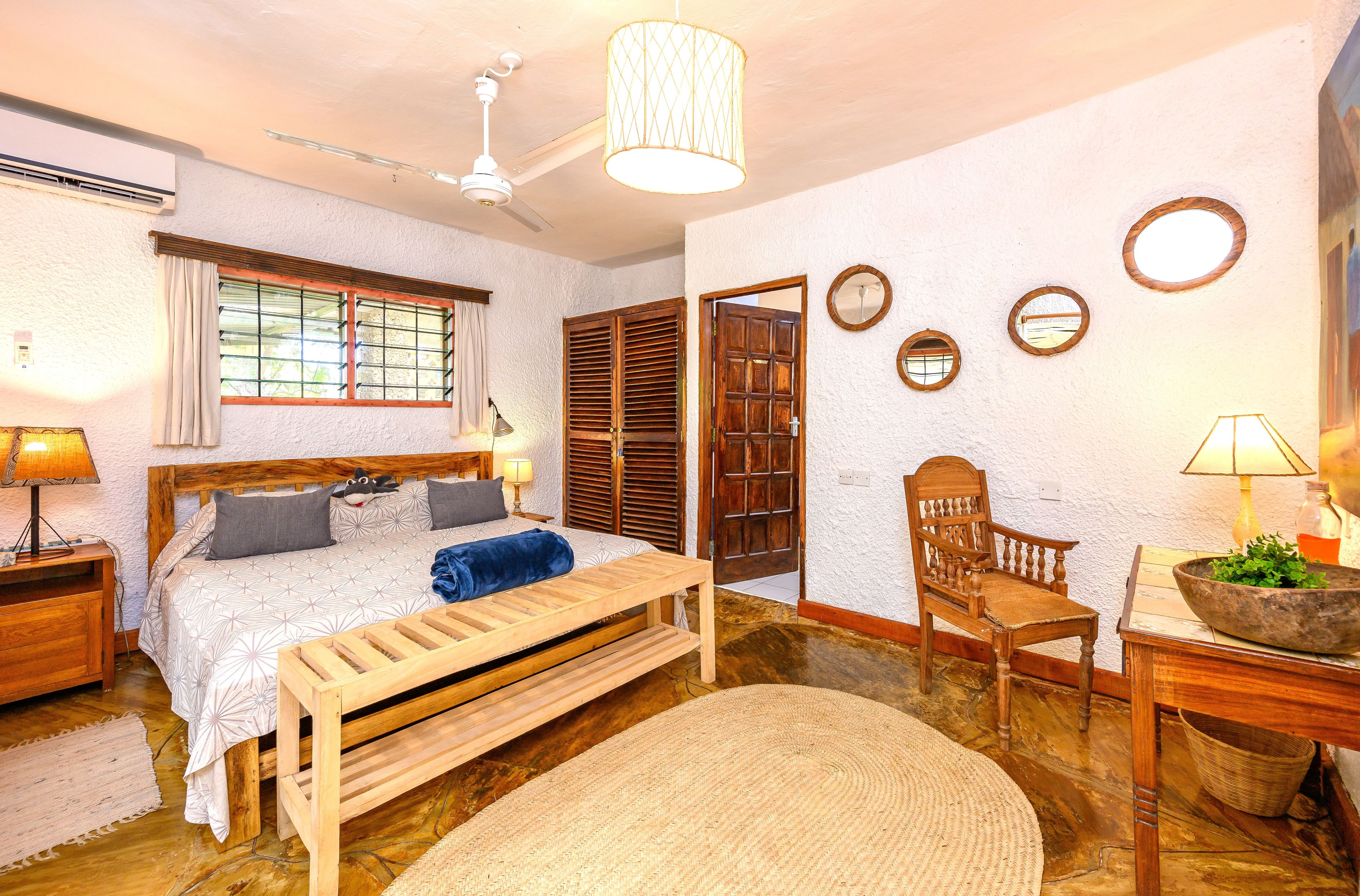 Une chambre à coucher comprenant un lit, des tables de chevet, un banc en bois, une chaise, une lampe, un ventilateur de plafond et une fenêtre avec des rideaux.
