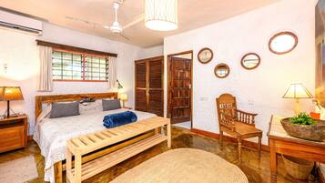Une chambre à coucher comprenant un lit, des tables de chevet, un banc en bois, une chaise, une lampe, un ventilateur de plafond et une fenêtre avec des rideaux.