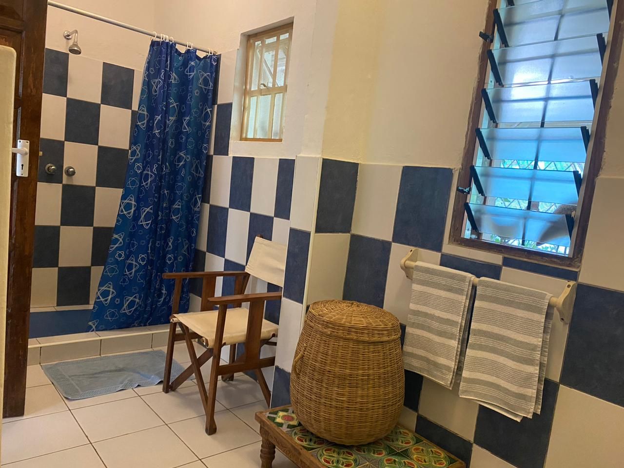A bathroom with a checkered tile wall, a shower with a blue curtain, a wooden chair, a woven basket, and a window with blinds.