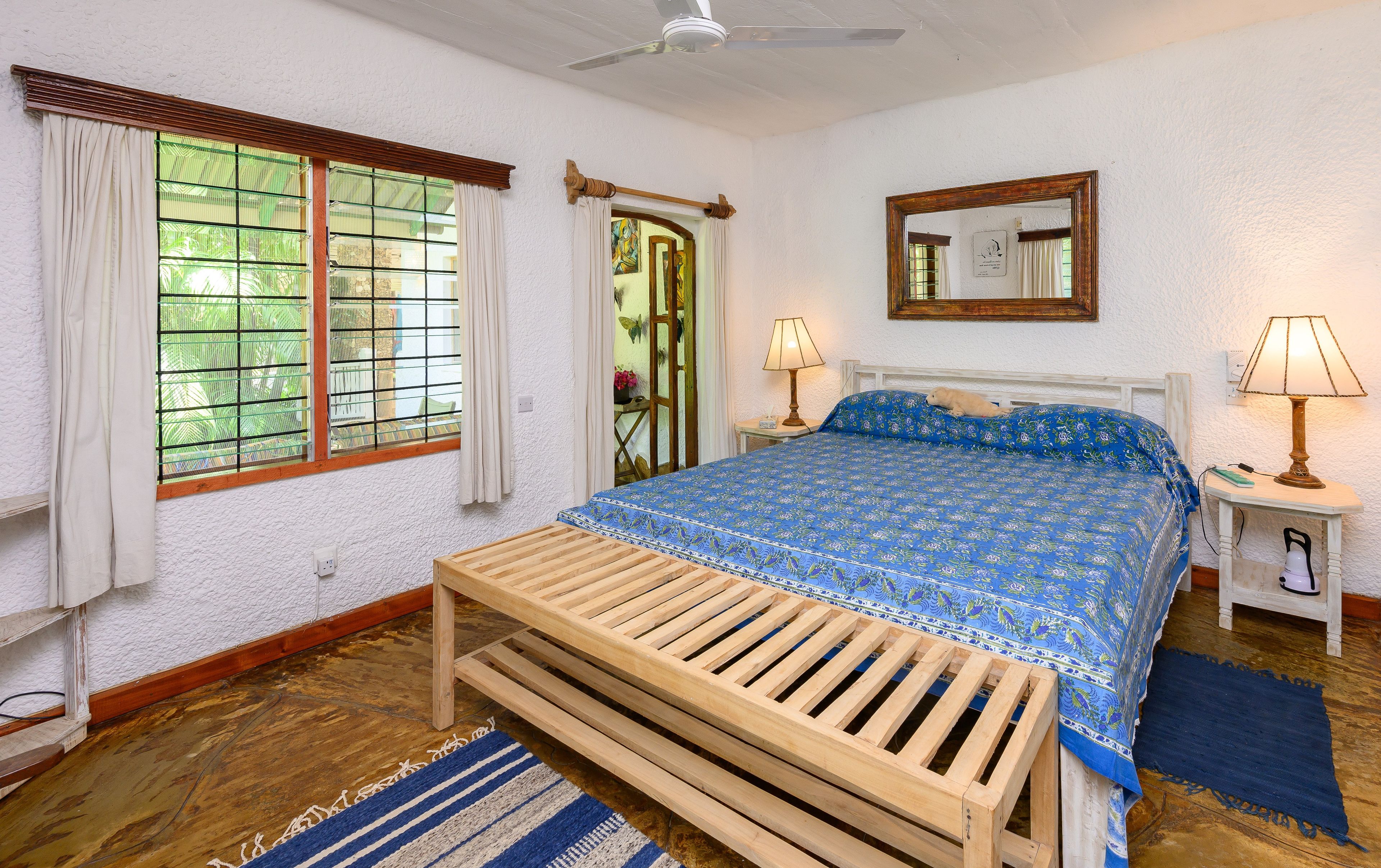 Un dormitorio con un banco de madera, una cama con colcha azul estampada, dos lámparas de noche, un espejo y una ventana con cortinas.