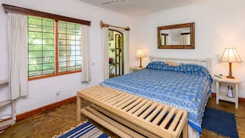 Un dormitorio con un banco de madera, una cama con colcha azul estampada, dos lámparas de noche, un espejo y una ventana con cortinas.
