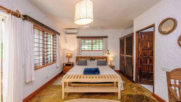 Une chambre à coucher comprenant un lit, des tables de chevet, une lampe, un luminaire suspendu, une fenêtre avec des rideaux et une porte avec un miroir.