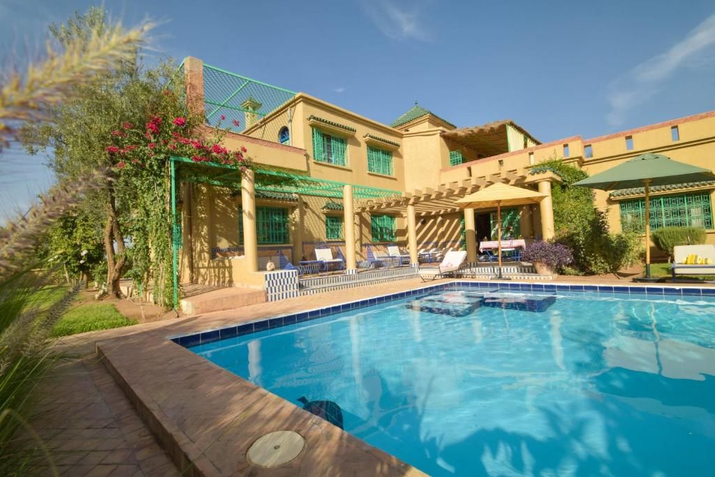 Una piscina con sillas de descanso y una pérgola, rodeada de vegetación y un edificio con múltiples ventanas.