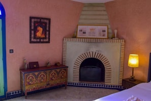 Une pièce avec une cheminée, un lit, une table de chevet avec une lampe et un meuble décoratif.