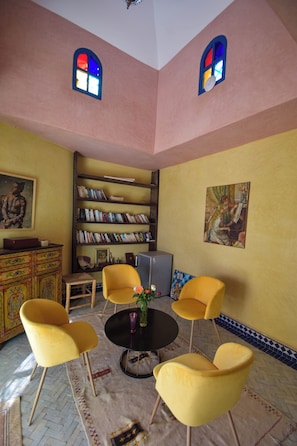 A room with a round table, four yellow chairs, a bookshelf, and two windows with stained glass.