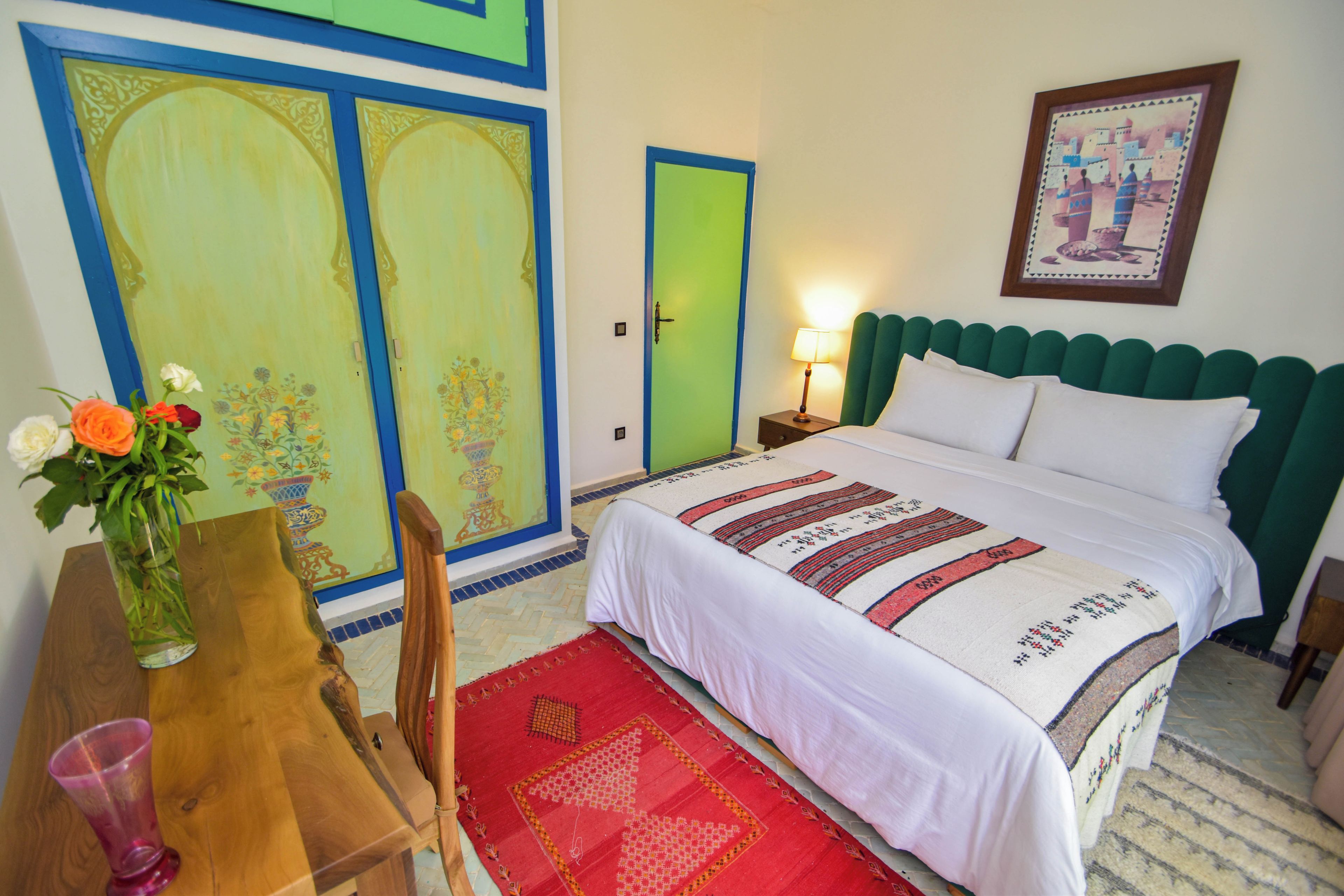 A hotel room with a bed, a nightstand, a lamp, a chair, a table with a vase of flowers, and two doors with decorative panels.