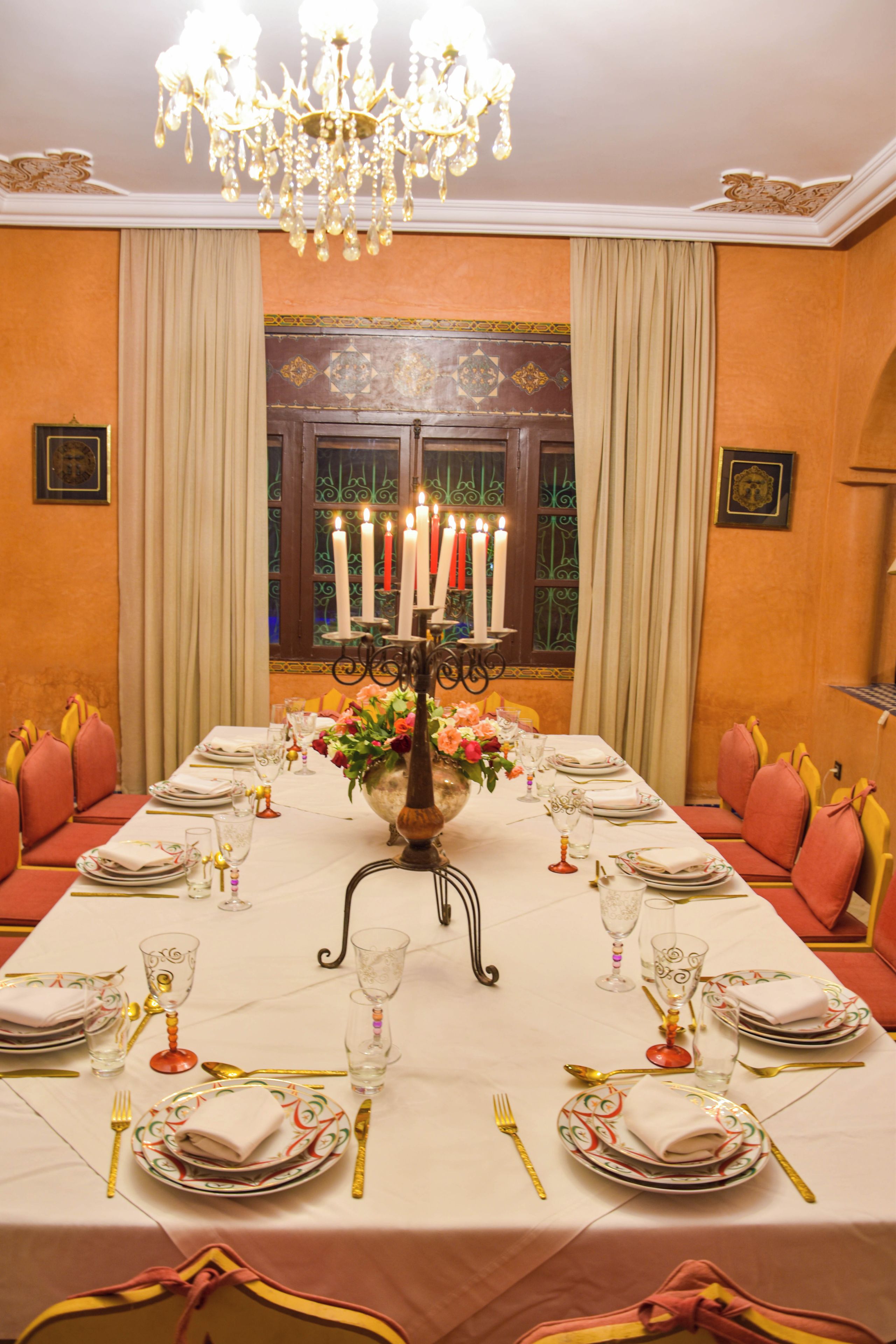 A formal dining room with a long table set for multiple guests, featuring a chandelier, candles, and a floral centerpiece.