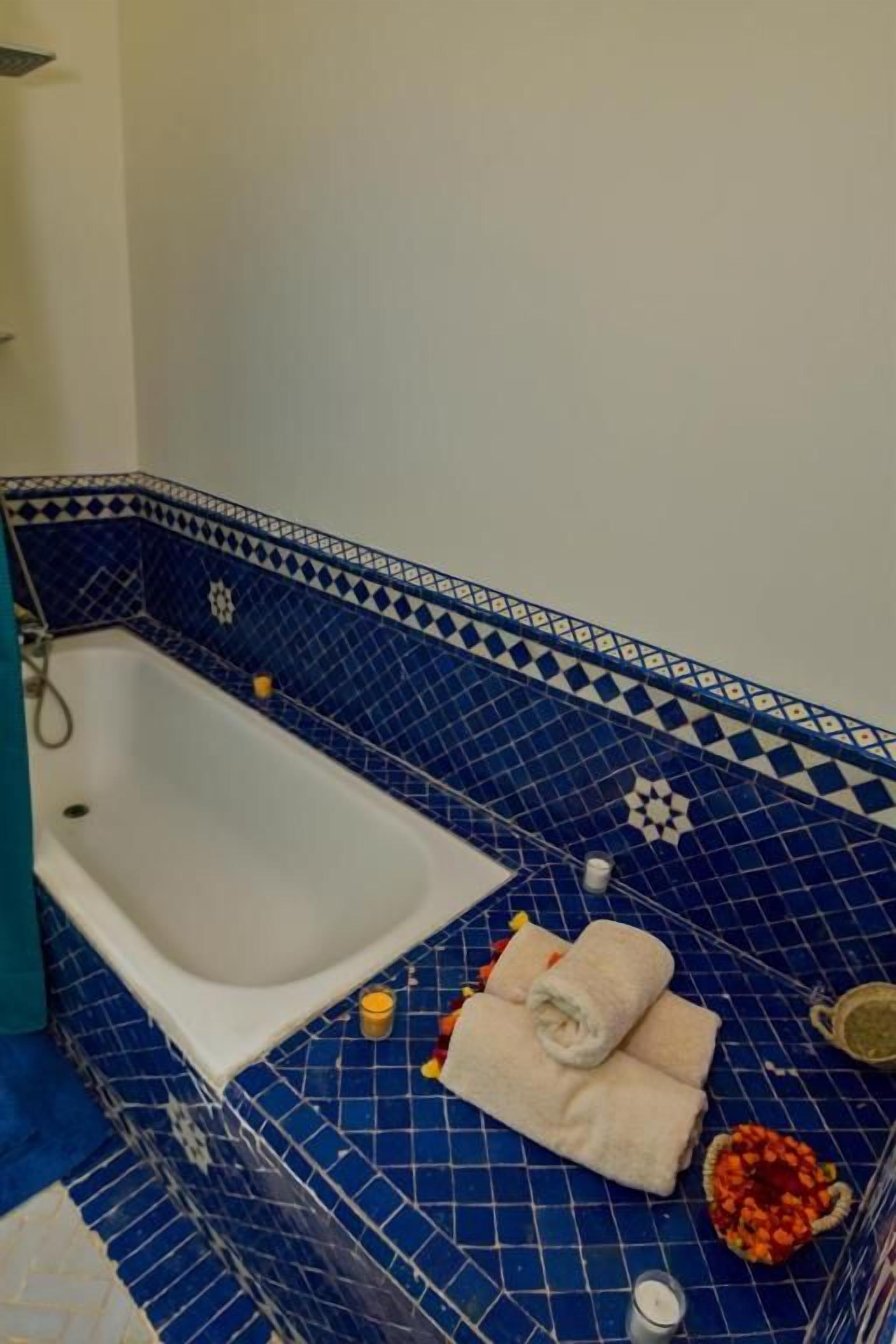 A bathroom with a white bathtub, blue patterned tiles, and a towel on the floor.
